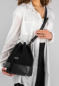 Femme portant une chemise blanche et un pantalon noir, tenant un sac seau en cuir et tissu noir par sa bandoulière sur l'épaule.