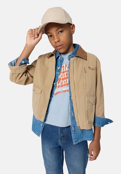 Garçon portant une casquette beige, une veste marron clair sur une chemise en denim bleue et un t-shirt graphique, ajustant sa casquette d'une main, debout devant un fond blanc.