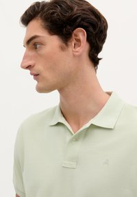 Polo de color verde claro hecho de tejido texturizado, con un cuello de tres botones y un pequeño diseño bordado en el pecho.