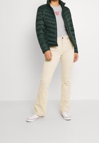 Vihreä puhvitakki, harmaa t-paita punaisella logolla, beige leveälahkeiset housut ja valkoiset tennarit. Takissa on vanutettu pinta ja korkea kaulus.