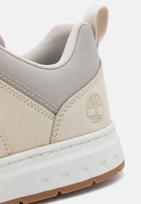 Beige Sneaker mit einer strukturierten Oberseite aus leichtem Material, weißer Sohle mit Rillen und geprägtem Logo-Detail an der Ferse.