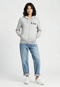 Haut de sweat à capuche zippé gris clair avec "sois gentil." brodé en noir, capuche doublée en beige, associé à un jean bleu à revers et des baskets blanches.