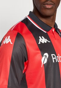 Κόκκινη και μαύρη ριγέ μπλούζα πόλο με υφή και μαύρο γιακά. Διαθέτει λευκά λογότυπα στους ώμους και το στήθος.