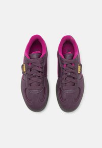 Chaussures de sport violettes avec des accents roses, présentant un matériau lisse, un design à lacets et une semelle en caoutchouc. Logo notable sur le côté.