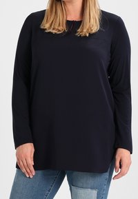 Námořnická modrá dlouhá rukávová blůza s kulatým výstřihem a mírně volným střihem. Měkká, hladká textura a minimalistický design. Sladěno s bledě modrými džíny.