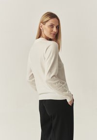 Pulover polo crem cu mânecă lungă, cu un guler clasic, textură moale și mansete răsfrânte, purtat împreună cu pantaloni negri cu picioare largi.