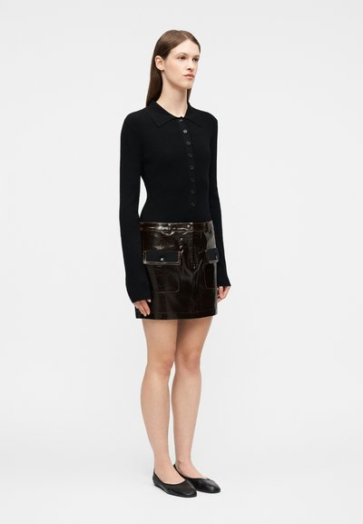 Γυναίκα στέκεται με το πρόσωπο προς τα εμπρός φορώντας μαύρο μακρυμάνικο πουκάμισο με κουμπιά, γυαλιστερό μαύρο μίνι φούστα με μπροστινές τσέπες και μαύρες μπαλαρίνες.