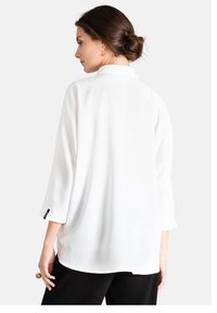 Blusa blanca con cuello, mangas tres cuartos y un corte relajado. Textura suave con dobladillo curvado; presenta una pequeña etiqueta negra en el puño.