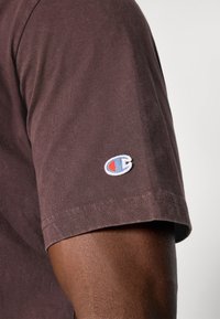 Braunes Baumwoll-T-Shirt mit kurzen Ärmeln; verfügt über ein kleines, weißes und blaues gesticktes Logo am linken Ärmel. Sanfte Textur und entspannte Passform.