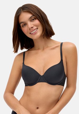 Mujer con cabello castaño hasta los hombros sonriendo, lleva un sujetador negro con alambre y tirantes finos ajustables, sobre un fondo claro y liso.