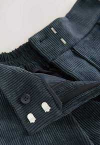 Pantalon en velours côtelé bleu foncé avec des boutons, des crochets en métal, une fermeture éclair et des détails de taille élastique sur un fond clair.