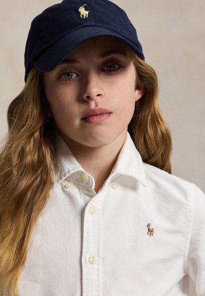 Persona joven con cabello largo que lleva una gorra azul marino y una camisa blanca abotonada con un pequeño logo bordado de un caballo.