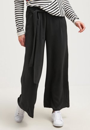 Czarne spodnie o szerokich nogawkach wykonane z gładkiego materiału, z wiązanym paskiem w talii i luźnym krojem, zestawione z białymi trampkami.