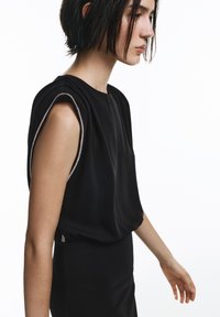 Črna majica brez rokavov z nabranim materialom, srebrnimi detajli ob šivih na rokah, mehko teksturo in sproščenim krojem.