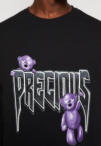 Maglietta nera con testo grigio crepato "PRECIOUS" e due orsetti viola con gli occhi sbarrati integrati nelle lettere.