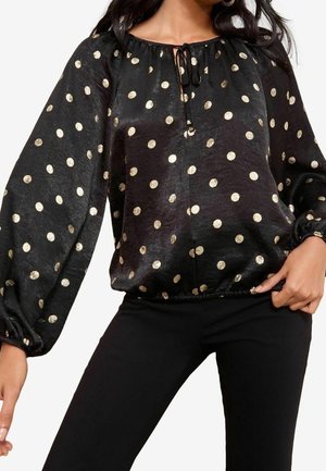 Blouse noire à manches longues avec des pois dorés, coupe ample, ourlet et poignets élastiqués froncés, portée avec un pantalon noir.