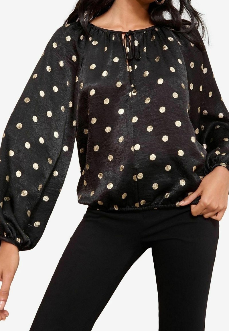 Blouse noire à manches longues avec des pois dorés, coupe ample, ourlet et poignets élastiqués froncés, portée avec un pantalon noir.