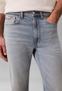 Άνδρας με ανοιχτού μπλε τζιν Calvin Klein, με το ένα χέρι στην τσέπη και λευκό πουκάμισο με μακριά μανίκια, μπαίνει μέσα στο παντελόνι.