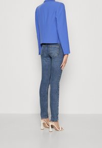 Blazer bleu cintré, jean en denim ajusté et talons blancs à brides. Le blazer est orné de boutons sur les manches ; le jean présente un classique design à cinq poches.