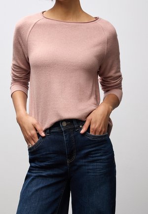 Vrouw met een lichtroze top met lange mouwen en donkerblauwe jeans met handen in de zakken tegen een effen grijze achtergrond.