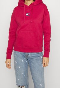 Červená mikina s kapucí a přední kapsou, se znakem Tommy Jeans, v kombinaci se světle modrými obnošenými džíny s bílými skvrnami od barvy.