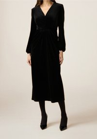 Abito midi in velluto nero con maniche lunghe, scollatura a V, corpino drappeggiato e un leggero luccichio, abbinato a eleganti tacchi neri e collant.