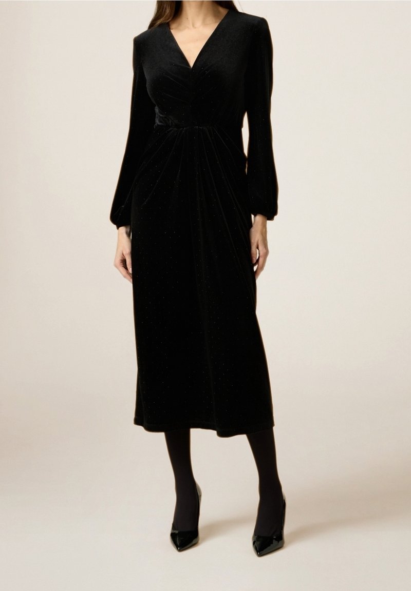 Abito midi in velluto nero con maniche lunghe, scollatura a V, corpino drappeggiato e un leggero luccichio, abbinato a eleganti tacchi neri e collant.