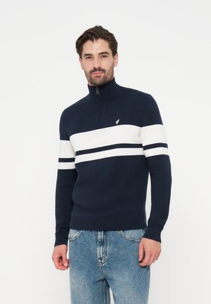 Mann trägt einen marineblau-weiß gestreiften Halb-Reißverschluss-Pullover und blaue Jeans, steht vor einem schlichten weißen Hintergrund.