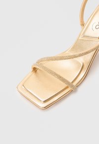 Gold metallic high-heel sandal with a square toe and two diagonal rhinestone-embellished straps, featuring a sleek, smooth texture.