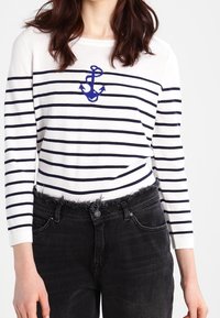 Wit katoenen shirt met lange mouwen en marineblauwe strepen, met een geborduurde anker. Gecombineerd met versleten zwarte jeans.