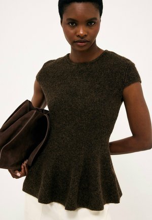 Donna con top peplum in maglia marrone a maniche corte che tiene una grande borsa marrone piegata su uno sfondo uniforme.