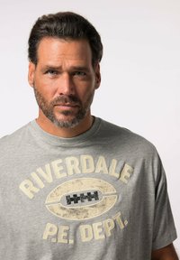 Camiseta gris de manga corta con el texto "DEPARTAMENTO DE EDUCACIÓN FÍSICA RIVERDALE" y un gráfico de fútbol vintage. Material suave con un corte casual y cuello redondo.