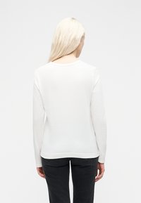 Femme aux longs cheveux blonds portant un t-shirt blanc à manches longues uni et un jean noir, vue de dos sur un fond blanc.