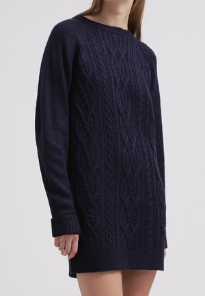 Robe-pull manches longues en maille torsadée bleu marine avec poignets et ourlet côtelés, portée par une personne sur un fond uni clair.