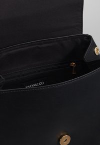 Sac à main en cuir noir avec une poche intérieure zippée, des éléments en métal doré et une texture lisse. Étiquette de marque visible à l'intérieur.