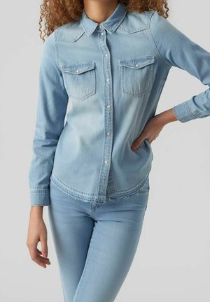 Femme portant une chemise en denim bleu clair et un jean, debout avec une main sur la hanche et l'autre détendue sur le côté.