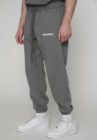 Graue Jogginghose mit elastischem Bund und Kordelzug, mit weißem "MULTIPLY"-Schriftzug, zulaufenden Beinen und gerippten Bündchen, kombiniert mit weißen Turnschuhen.
