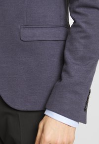 Tmavě modrý strukturovaný blazer s úzkým střihem, který má náprsní kapsu a dekorativní knoflíky na manžetách. Viditelná manžeta světle modré košile.