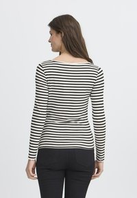 O femeie cu păr lung și brunet, îmbrăcată cu un tricou cu mâneci lungi, cu dungi orizontale alb-negru și pantaloni negri, întorsă cu spatele pe un fundal alb.
