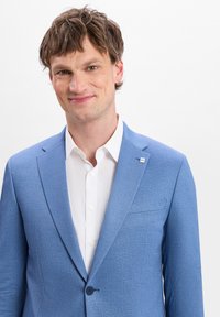 Hellblau strukturierter Blazer mit Revers, einem einzelnen Knopfverschluss und einem weißen Hemd. Enthält ein kleines, markiertes Detail am Revers.
