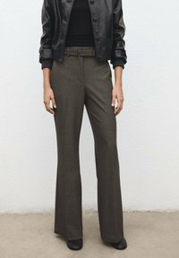 Kobieta w szerokich spodniach z szerokim paskiem w kolorze ciemnoszarym, czarnym dopasowanym golfie, czarnej skórzanej kurtce i czarnych butach, stojąca na tle białej ściany.