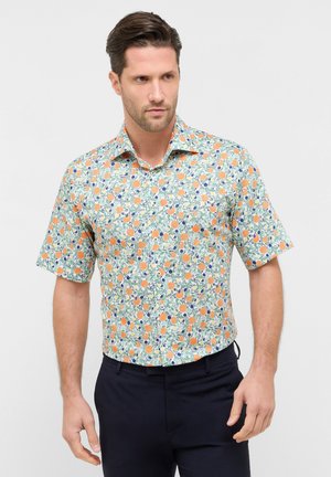 Mann trägt kurzärmeliges Hemd mit orange-grünem Obst- und Blattmuster, kombiniert mit dunkler Hose, steht vor einfarbigem Hintergrund.