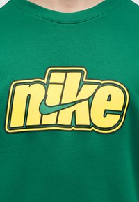 Πράσινο μπλουζάκι με μεγάλο κίτρινο λογότυπο "nike" και μαύρο περίγραμμα και σχέδιο swoosh στο κέντρο του στήθους.