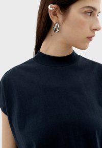 Schwarzes Oberteil mit hohem Kragen aus weichem Stoff, mit kurzen Ärmeln. Silberne Creolen und ein dekorativer Ohrcuff betonen den Look.