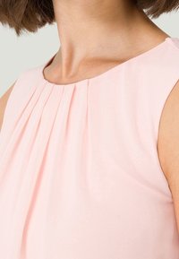 Blusa sin mangas de color rosa claro con pliegues sutiles en el escote, hecha de una tela suave, que presenta un diseño simple y elegante.