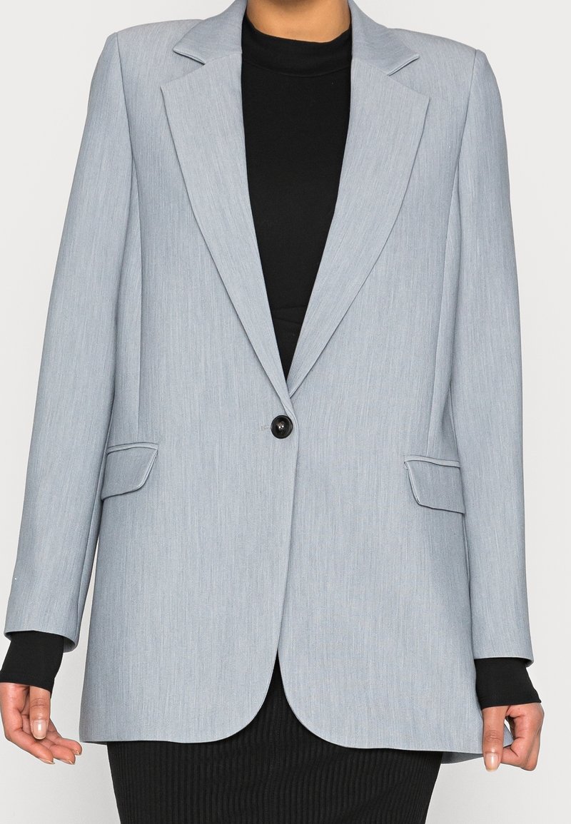 Blazer gris clair à un bouton unique avec revers crantés porté sur un top noir à manches longues et une jupe noire côtelée.