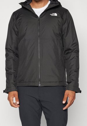 Mann trägt eine schwarze The North Face Jacke mit Reißverschluss und Kapuze, kombiniert mit schwarzer Hose, stehend vor einem schlichten hellen Hintergrund.