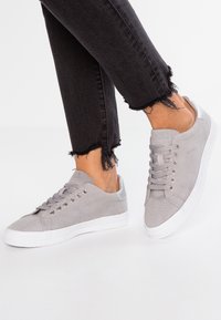 Szare zamszowe sneakersy mają białą teksturowaną gumową podeszwę, metaliczne akcenty oraz zaokrąglony czubek. Noszone z postrzępionymi czarnymi dżinsami.