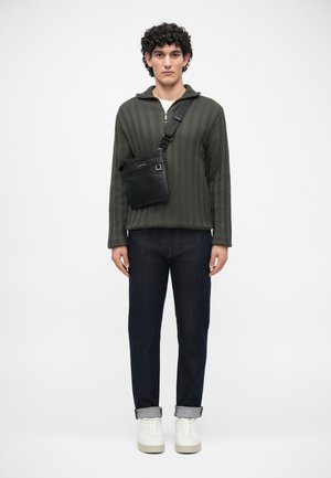 Muž stojící ve tmavě zeleném strukturovaném svetr s zipem, tmavých manžetových džínách, bílých teniskách a černé crossbody tašce na pozadí s čistě bílým pozadím.