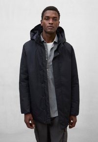 Czarna, wodoodporna parka z kapturem, wyposażona w przedni zamek błyskawiczny i zatrzaski, noszona na jaśniejszej, szarej koszuli, na jednolitym tle.